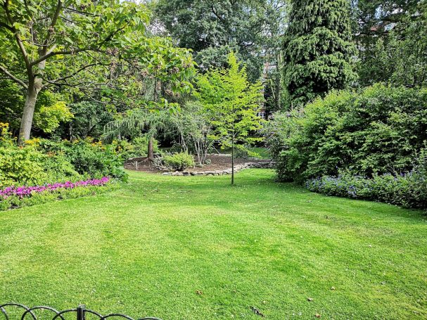 Grüner Garten mit Blumenbeeten, Rasenfläche und Bäumen im Hintergrund.