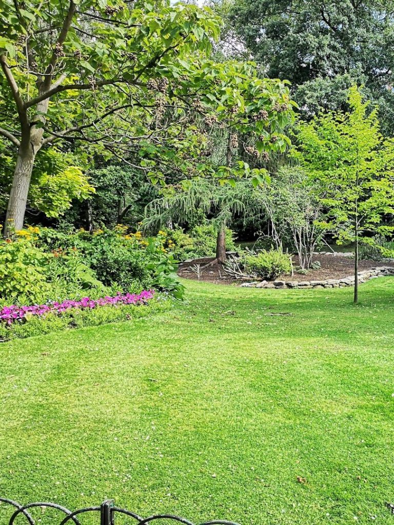 Grüner Garten mit bunten Blumenbeeten und einem kleinen Teich im Hintergrund.