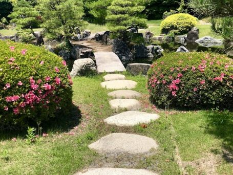 Steinerner Weg durch einen Garten mit blühenden Sträuchern und Brücke über einen Teich.
