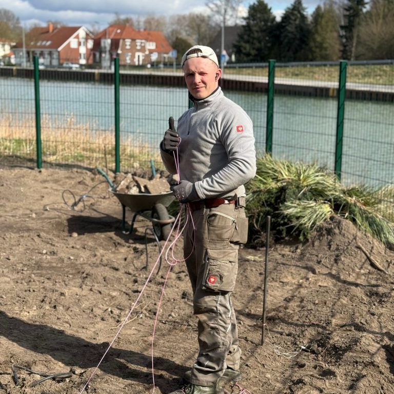 Mann mit Werkzeug steht an einer Baustelle nahe einem Gewässer.